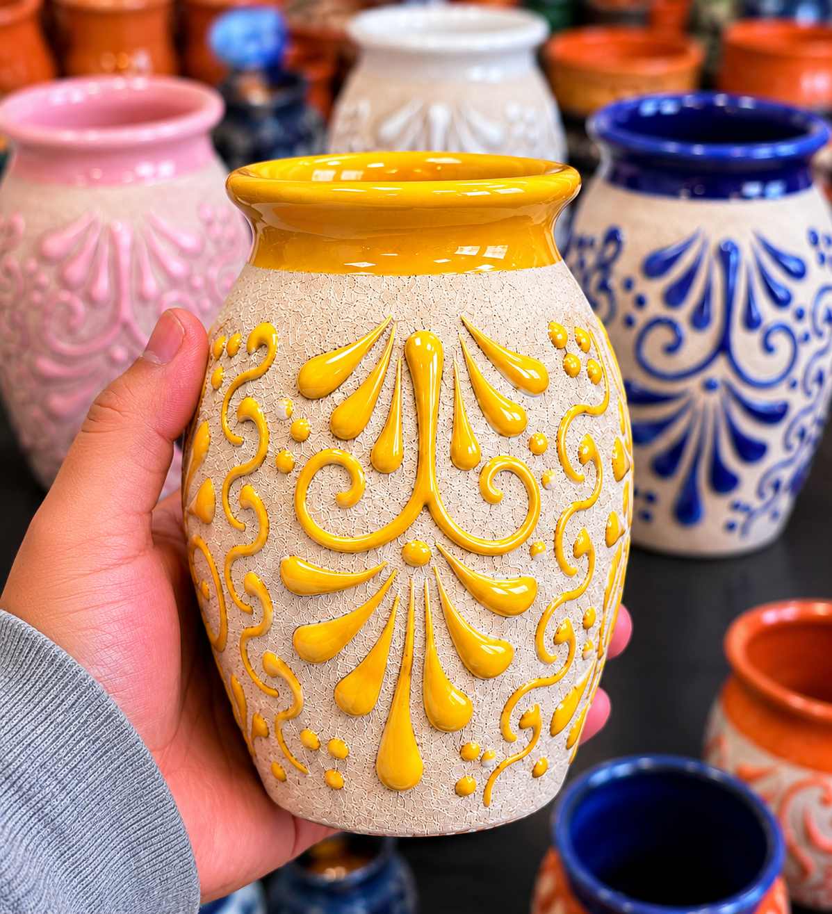 Hand holding a ceramic vase with yellow patterns against a background of various colorful vases.